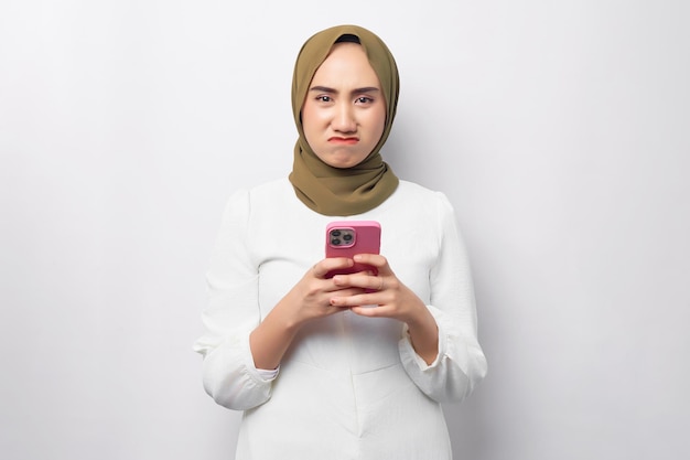Linda e triste mulher muçulmana asiática usando hijab segurando o celular na mão e olhando a câmera isolada no retrato de estúdio de fundo branco Pessoas conceito de estilo de vida religioso