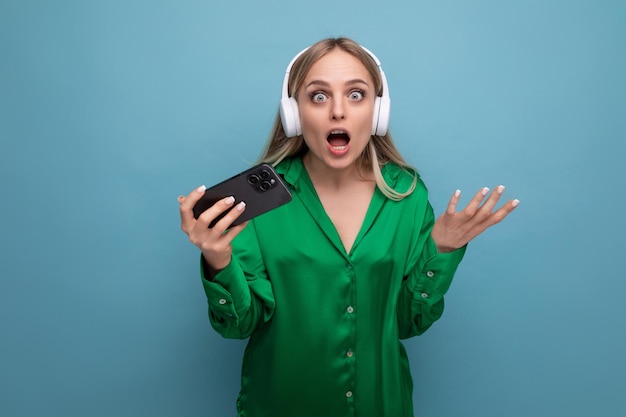 Linda emocionada mujer joven rubia atractiva sorprendida con auriculares y teléfono inteligente sobre fondo azul