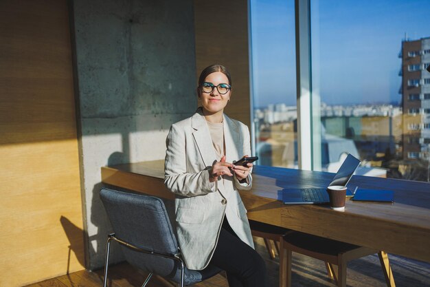 Linda empleada positiva con cabello largo y rubio en ropa informal mirando el teléfono mientras trabaja en un nuevo proyecto de negocios en la mesa con una computadora portátil y aparatos en la oficina