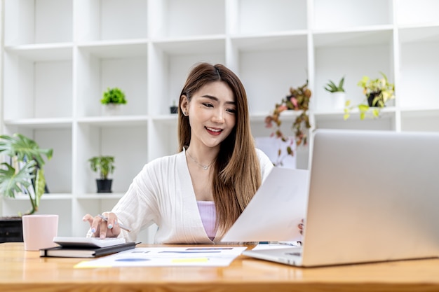 Linda empresária asiática sentada em seu escritório particular, conversando com seu parceiro via laptop e verificando documentos, ela é uma executiva de uma empresa iniciante. Conceito de gestão financeira.