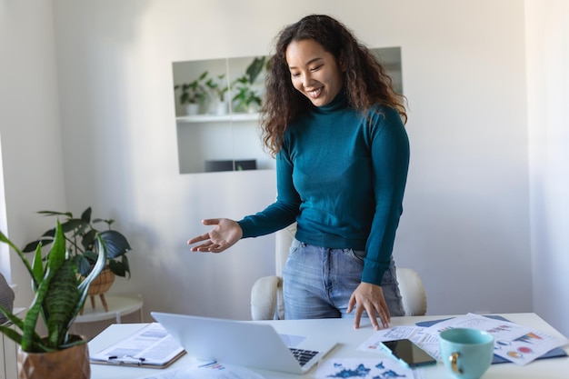 Linda empresária asiática sorridente lendo algo em um laptop enquanto se inclina sobre a mesa no escritório