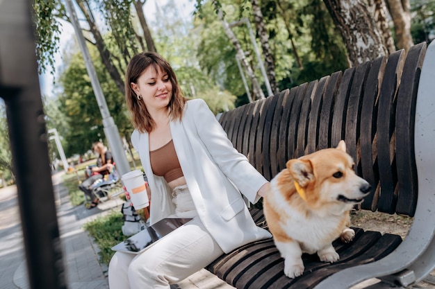 Linda empresária de terno branco sentada no parque da cidade bebe café e trabalhando em tablet digital com seu cachorro Welsh Corgi Pembroke