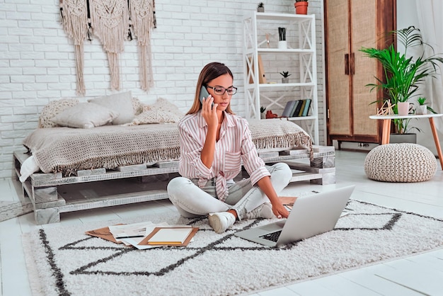 Linda empresária em roupas casuais e óculos está usando um laptop falando no celular