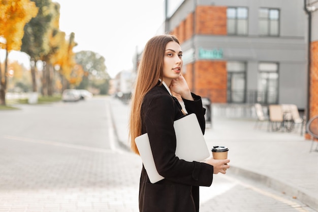 Linda empresária profissional de sucesso em um elegante casaco preto com um laptop e um café andando na cidade xA