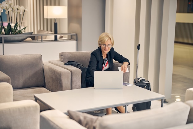 Linda empresária sentada à mesa com o notebook e segurando o smartphone enquanto espera seu voo no corredor