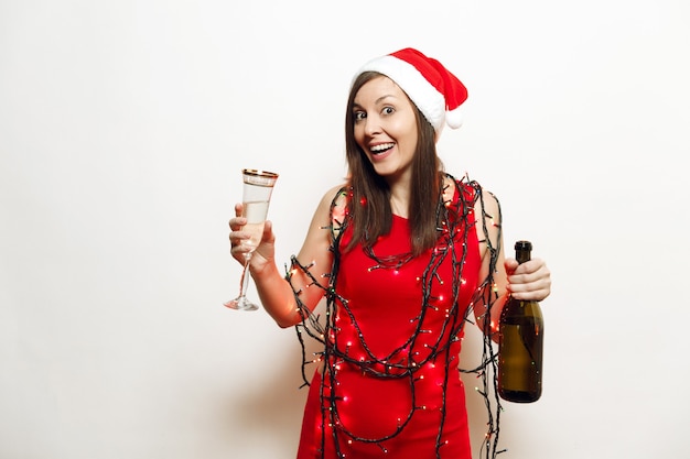 Linda encantadora jovem sorridente feliz mulher com vestido vermelho, chapéu de Natal, lanternas de guirlanda em pé com copo e garrafa de champanhe no fundo branco. Santa menina isolada. Feriado de ano novo de 2018.