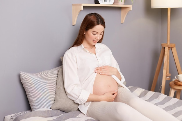 Linda encantadora mujer caucásica de cabello castaño embarazada con camisa blanca sentada en la cama tocando sosteniendo su vientre relajándose descansando en los últimos meses de embarazo