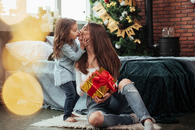 Linda escena de beso. Madre e hija se sientan en una habitación decorada de vacaciones y tiene una caja de regalo