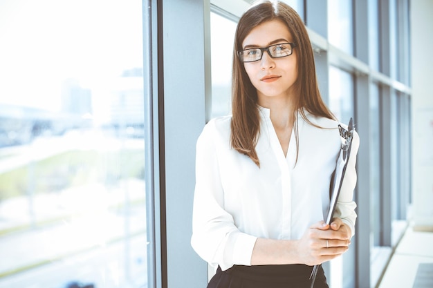 Linda especialista feminina com área de transferência em pé no escritório moderno e sorrindo encantadoramente Trabalhando na estratégia de plano de análise de dados de design Conceito de pessoas de negócios