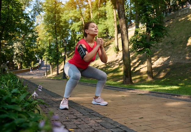 Linda esportista confiante fazendo agachamentos profundos desfrutando de exercícios ao ar livre no dia quente e ensolarado de verão Esporte fitness treinamento funcional com peso corporal