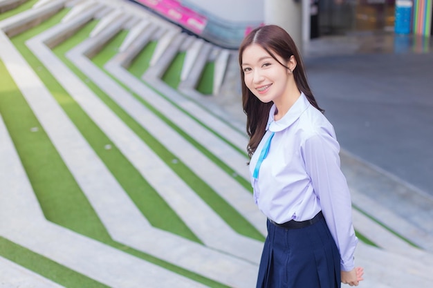 Linda estudante asiática do ensino médio no uniforme escolar fica e sorri alegremente.