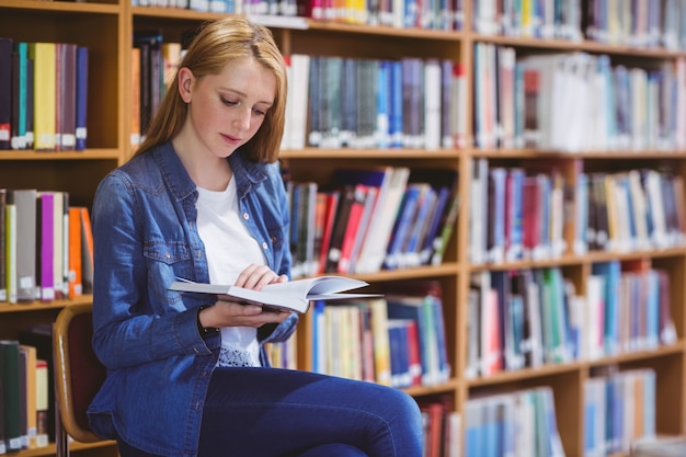 Linda estudiante sentada en la silla leyendo el libro en la biblioteca