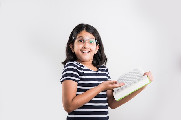 Linda y estudiosa niña india o asiática sosteniendo o estudiando o leyendo un libro mientras está de pie aislado sobre fondo blanco.