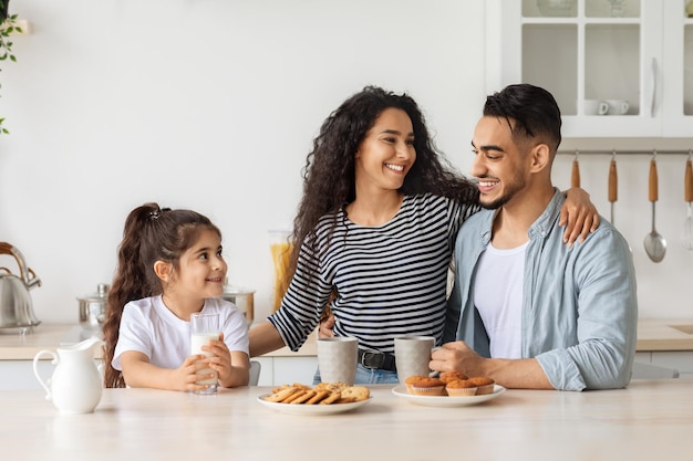 Foto linda familia árabe feliz tomando un refrigerio mientras pasan el fin de semana juntos en casa, alegre padre joven, madre y niña rizada sentada en la cocina, bebiendo café y leche con galletas caseras