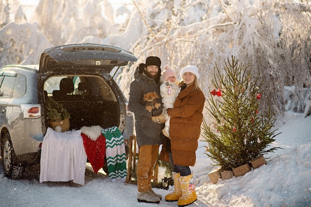 Linda família em pé ao lado do porta-malas de um SUV na floresta de inverno, comemorando o Natal