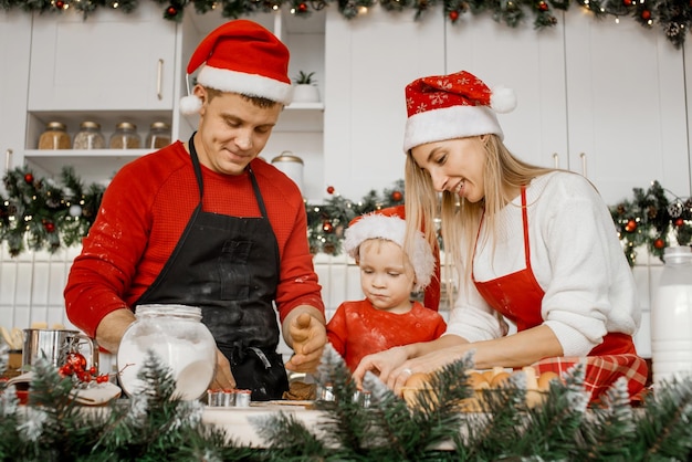 Linda família jovem em chapéus de Papai Noel se divertindo e amassando a massa enquanto assava biscoitos de Natal