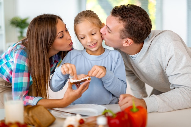 Linda família jovem preparando uma refeição saudável para o café da manhã na cozinha doméstica. A mãe aprende a menina a fazer o sanduíche e o pai está beijando a filha.