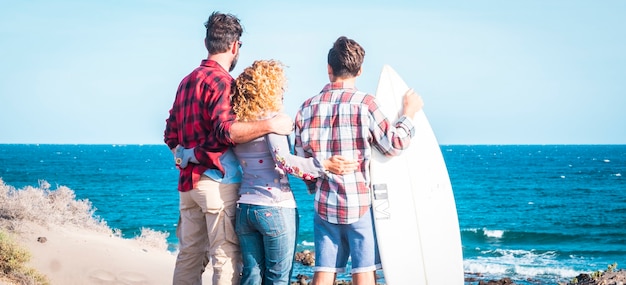 Linda família reunida para sempre na praia com um dia de sol e um adolescente surfista - conversando com a mesa de surf e pessoas felizes - férias de verão - vista traseira - família feliz abraçada