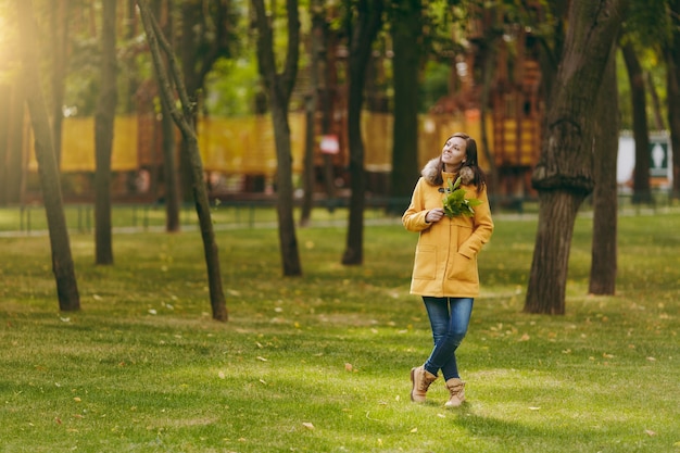 Linda feliz caucasiana jovem sorridente de cabelo castanho no casaco amarelo, jeans, botas na floresta verde. Moda modelo feminino com folhas douradas de outono em pé e andando no parque do início do outono ao ar livre.