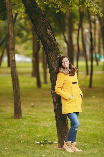 Linda feliz caucasiana jovem sorridente de cabelo castanho no casaco amarelo, jeans, botas na floresta verde. Moda modelo feminino com folhas douradas de outono em pé e andando no parque do início do outono ao ar livre.