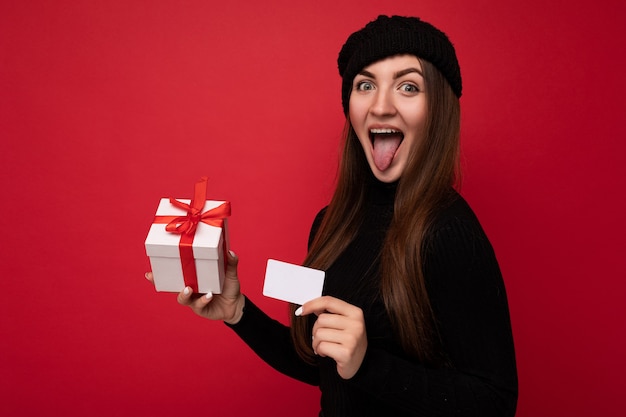 Linda feliz engraçada jovem morena vestindo um suéter preto e um chapéu isolado em um fundo vermelho