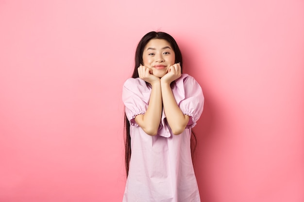 Linda y feliz mujer asiática esperando algo con entusiasmo, con la cara inclinada en las manos y mirando la hermosa escena con admiración, de pie contra el fondo rosa.