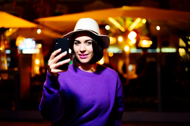 Linda feliz sorridente jovem fazendo selfie foto na rua à noite. Modelo olhando para a câmera, usando um chapéu e um suéter roxo