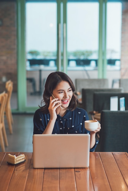 Linda fofa jovem empresária asiática no café, ouvindo o telefone celular e segurando uma caneca de café