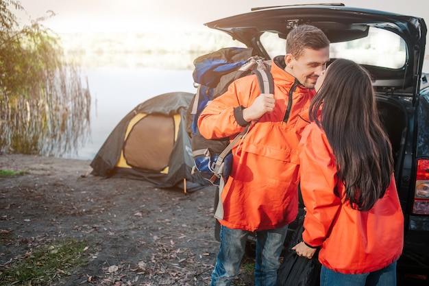 Linda foto do jovem casal a beijar. Eles usam jaquetas laranja. Pessoas em pé perto um do outro. Ele tem mochila. Casal está no tronco aberto e tenda.