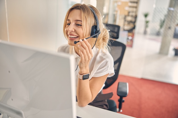 Linda funcionária de call center tocando o microfone do headset e sorrindo enquanto usa um computador moderno