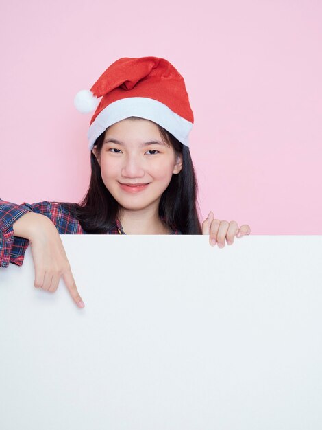 Linda garota adolescente com chapéu de Papai Noel, apontando o dedo em um cartaz em branco em pé no fundo rosa.