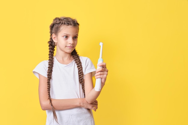 Linda garota adolescente com penteado trançado segurando uma escova de dentes elétrica nas mãos em um fundo amarelo, seleção de produtos de higiene bucal para crianças