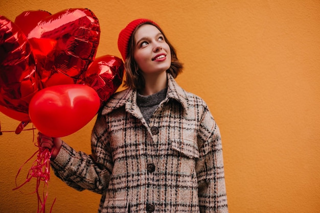 Linda garota adolescente de chapéu vermelho e jaqueta de tweed sonhadoramente posando com balões vermelhos contra o fundo da parede laranja