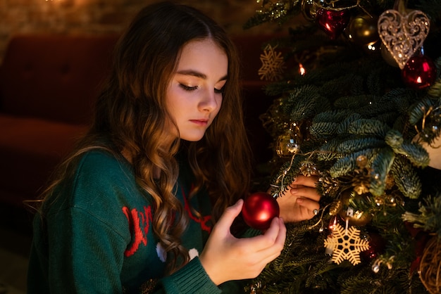 Linda garota adolescente decora a árvore de Natal com bolas vermelhas no suéter verde com veados aconchegantes e mágicos.