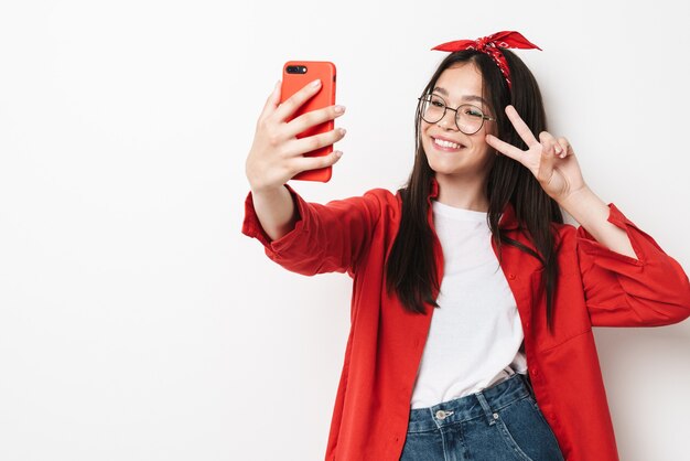 Linda garota adolescente feliz usando roupa casual em pé, isolada na parede branca, tirando uma selfie, gesto de paz