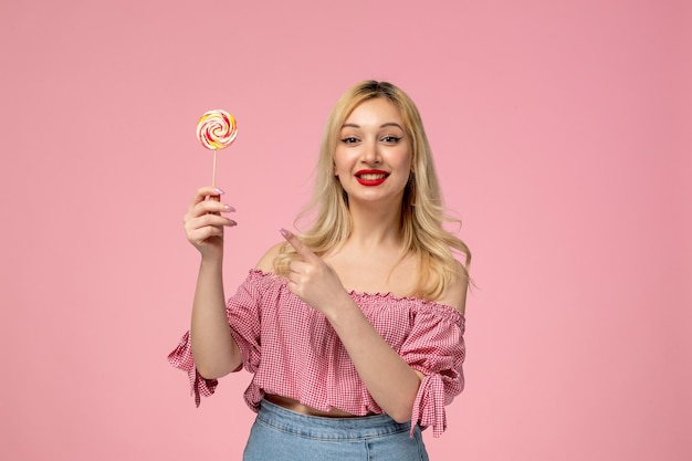 Linda garota adorável jovem com batom vermelho na blusa rosa apontando para pirulito alegremente