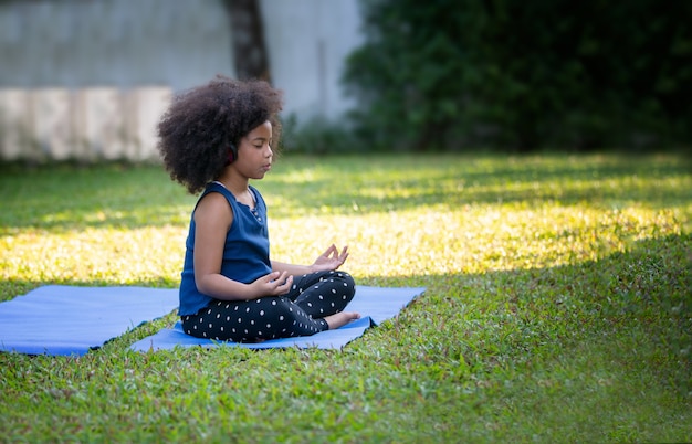 Linda garota africana com cabelo afro, fazendo meditação no tapete de ioga no jardim.