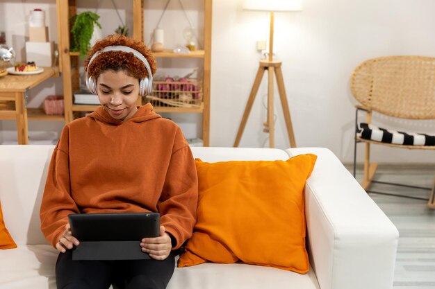 Linda garota afro-americana com penteado afro usando fones de ouvido sorrindo ouvindo música em casa interior Jovem gosta de ouvir música favorita no tablet Conceito de alegria de estilo de vida de pessoas