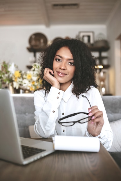 Linda garota afro-americana sentada no restaurante com o laptop e óculos na mão. Menina bonita trabalhando em um café com notebook e laptop na mesa