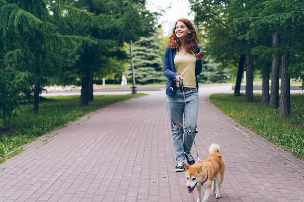 Linda garota andando com cachorro no parque verde segurando telefone inteligente sorrindo