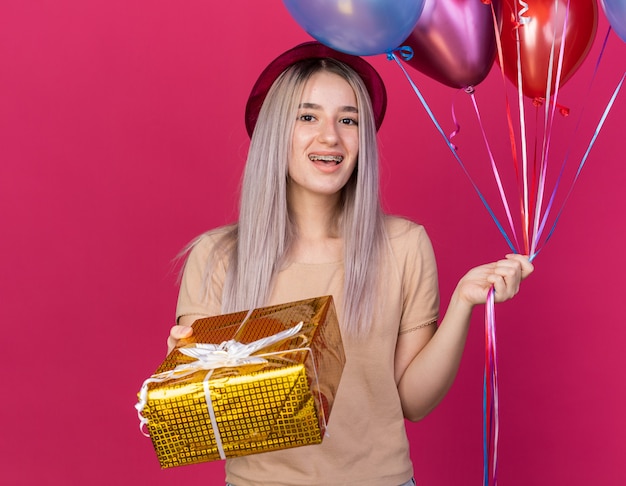 Linda garota animada usando chapéu de festa com aparelho dentário segurando balões com caixa de presente