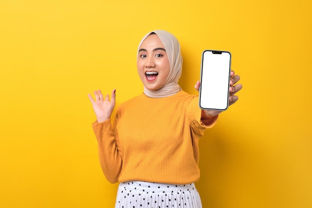 Linda garota asiática animada usando hijab mostrando celular com tela branca em branco levantando a mão dizendo sim comemorando boa sorte isolada no espaço de cópia de maquete de fundo amarelo