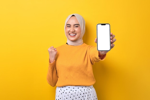 Linda garota asiática animada usando hijab mostrando celular com tela branca em branco levantando o punho comemorando boa sorte isolada no espaço de cópia de maquete de fundo amarelo