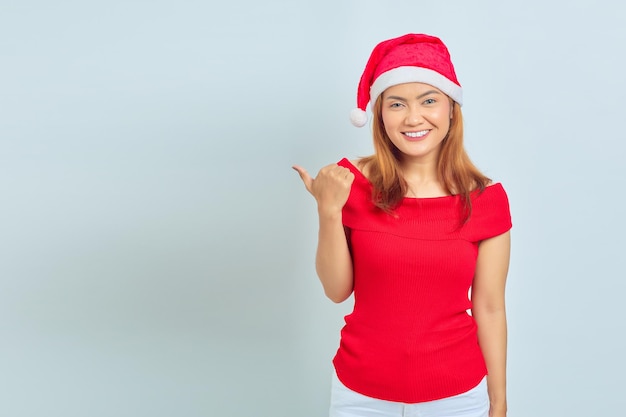 Linda garota asiática com chapéu de Natal com expressão facial sorridente e apontando para o lado