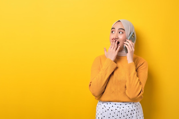 Linda garota asiática espantada usando hijab falando no celular com um amigo olhando para o espaço vazio isolado em fundo amarelo