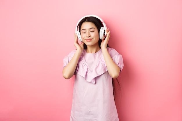 Linda garota asiática fecha os olhos enquanto ouve música em fones de ouvido, desfrutando de um som suave, em pé contra um fundo rosa.