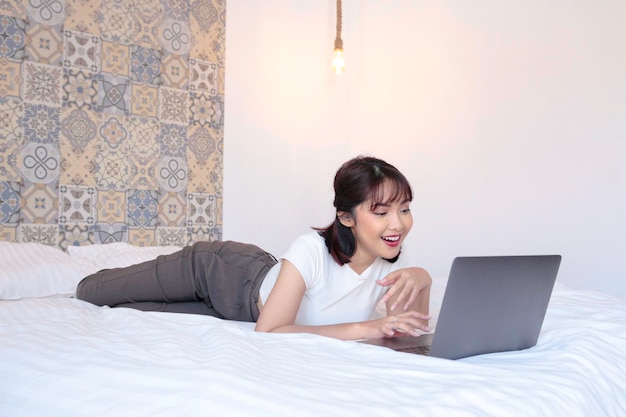 Linda garota asiática feliz e sorridente trabalha no laptop na cama Trabalha em casa conceito