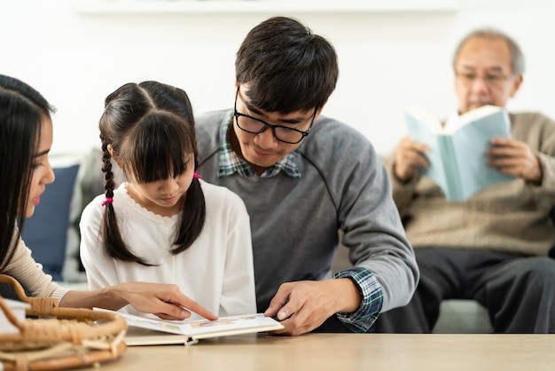 Linda garota asiática lendo um livro de desenho animado com a mãe e o pai na sala de estar