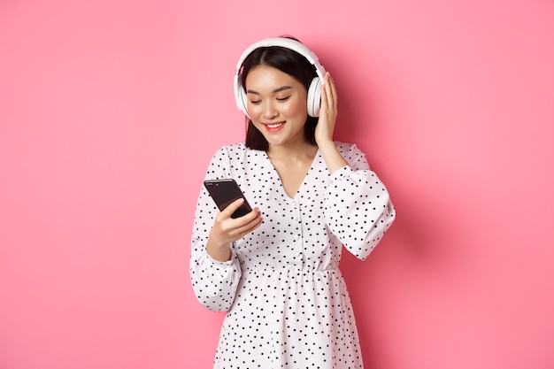 Linda garota asiática ouvindo música em fones de ouvido, olhando para o celular e sorrindo, em um vestido contra rosa.