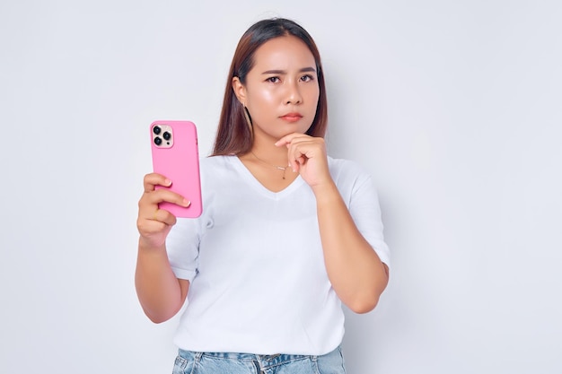 Linda garota asiática pensativa vestindo camiseta branca casual segurando o telefone celular rosto sério pensando perguntas isoladas no fundo branco Pessoas conceito de estilo de vida